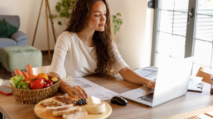 This image shows a woman with long, brown, curly hair sat at a wooden dining table in what appears to be a combined living room and dining room. She is sat opposite a laptop aith a bowl of fresh vegetables in a wicker basket to her right and a circular wooden chopping board behind it, with sliced bread, cheese and nuts on. She has some paperwork and clipboards filling the remaining space on the table and is looking intently at the latpop screen.